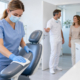 Eine Mitarbeiterin desinfiziert mit Handschuhen und Mundschutz die Armlehne eines Behandlungsstuhls in einer hellen Zahnarztpraxis. Im Hintergrund begrüßt ein Zahnarzt eine Patientin. Das Bild veranschaulicht sorgfältige Hygiene und eine vertrauensvolle Atmosphäre.