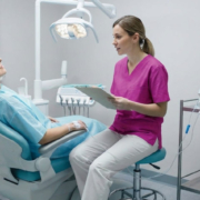 Das Bild zeigt eine Behandlungssituation in einer modernen Zahnarztpraxis. Eine Patientin liegt entspannt in einem Zahnarztstuhl und trägt einen Behandlungsumhang. An ihrem Arm ist ein intravenöser Zugang gelegt, der auf eine Sedierung oder medizinische Überwachung hinweist. Neben ihr sitzt eine medizinische Fachkraft in pinkfarbener Arbeitskleidung auf einem Hocker und hält ein Klemmbrett in der Hand, während sie die Patientin aufmerksam beobachtet oder mit ihr spricht. Im Hintergrund befinden sich typische Geräte einer Zahnarztpraxis: eine Behandlungsleuchte über dem Stuhl, ein Instrumentenarm sowie ein Monitor mit Vitaldatenanzeige. Die Szene vermittelt eine ruhige, kontrollierte Behandlungssituation, möglicherweise im Zusammenhang mit einer IV-Sedierung (Dämmerschlaf)