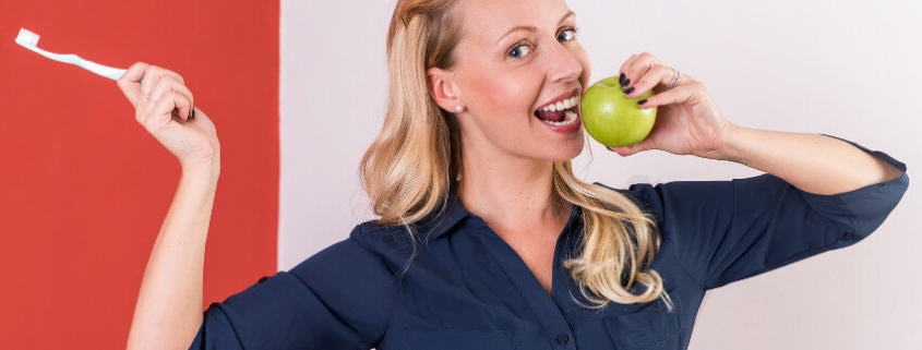 Frau mit Zahnbürste in der einen Hand und grünem Apfel in der anderen, lächelnd und zahnfreundliche Ernährung symbolisierend.
