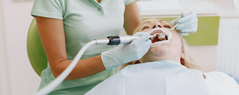 Zahnärztin in einem hellen Behandlungsraum, die mit Handschuhen und einem zahnärztlichen Instrument die Zähne einer liegenden Patientin behandelt