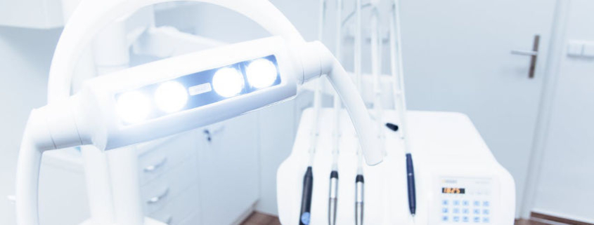 Das Bild zeigt eine moderne zahnärztliche Behandlungseinheit in einer hellen, klinisch sauberen Zahnarztpraxis. Im Vordergrund ist eine helle Untersuchungsleuchte mit mehreren LED-Lampen zu sehen, die auf den Behandlungsstuhl gerichtet ist. Im Hintergrund befinden sich mehrere zahnärztliche Instrumente, die ordentlich an einem Gerätehalter angebracht sind. Die Einrichtung ist in Weißtönen gehalten und vermittelt einen sterilen, professionellen Eindruck.