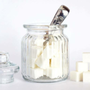 Ein Glasbehälter mit silbernem Löffel, gefüllt mit weißen Zuckerwürfeln, daneben ein stapel zusätzlicher Zuckerwürfel und der Glasdeckel des Behälters.