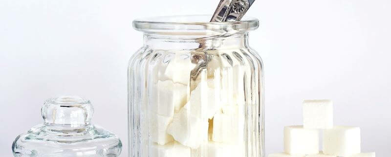 Ein Glasbehälter mit silbernem Löffel, gefüllt mit weißen Zuckerwürfeln, daneben ein stapel zusätzlicher Zuckerwürfel und der Glasdeckel des Behälters.
