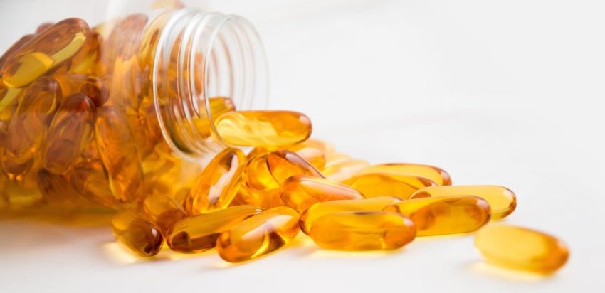 Ein Glasbehälter mit verschütteten goldfarbenen Vitamin-D3-Softgel-Kapseln auf weißem Hintergrund