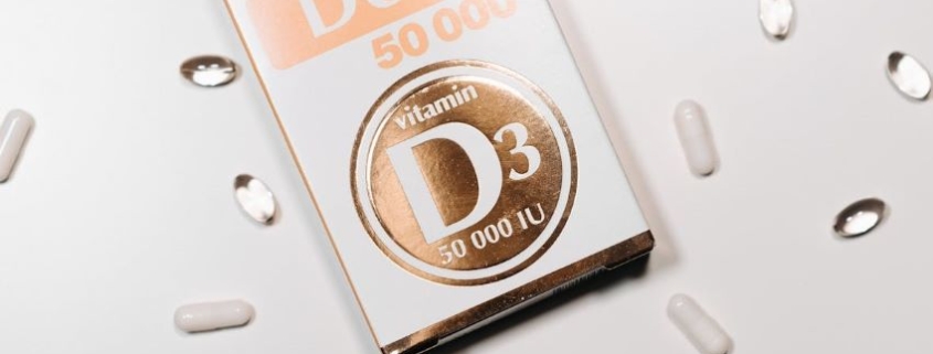 Verpackung von Vitamin D3 50.000 IU Cholecalciferol-Softgel-Kapseln, umgeben von weißen und durchsichtigen Kapseln auf weißem Hintergrund.