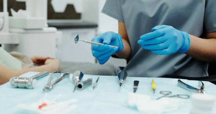 Ein Zahnarzt mit blauen Handschuhen hält ein Zahnspiegel, bereit für eine Untersuchung oder Behandlung, mit verschiedenen zahnärztlichen chirurgischen Instrumenten und Materialien auf einem Tablett im Vordergrund als Vorbereitung für eine Weisheitszahn-Entfernung. Der Patient ist außerhalb des Fokus, und der Hintergrund zeigt einen Teil des zahnmedizinischen Behandlungsstuhls und der Praxisumgebung
