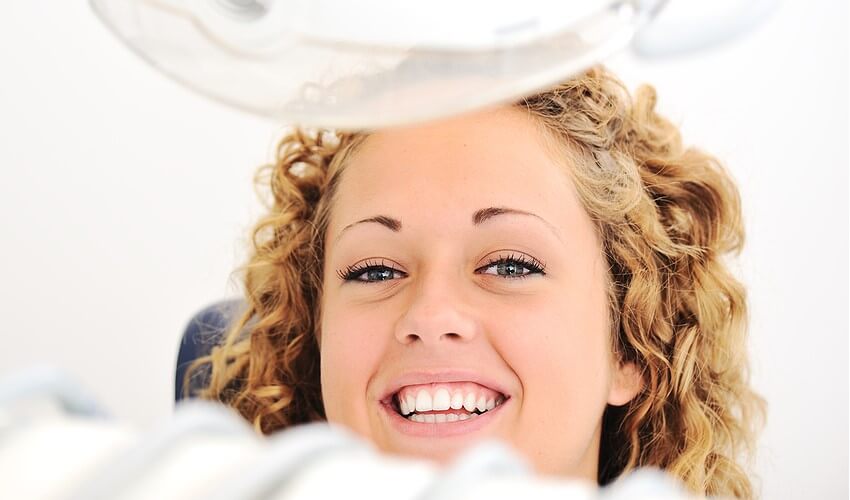 Eine junge Frau lächelt im Behandlungsstuhl beim Zahnarzt. Im Vordergrund ist ein Teil einer zahnärztlichen OP-Lampe und Teile von zahnärztlichem Instrumentarium zu sehen. Die Farben sind hell und freundlich und das Lächeln der frau symbolisiert Vitalität und Freude.