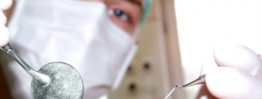 Patientenperspektive eines Zahnarztbesuches: Eine Person mit einer chirurgischen Maske und Schutzbrille hält zahnärztliche Instrumente – einen Spiegel und eine Sonde – in der Nähe der Kamera, was den Eindruck erweckt, als würde der Betrachter auf dem Zahnarztstuhl sitzen und auf den Beginn der Behandlung warten
