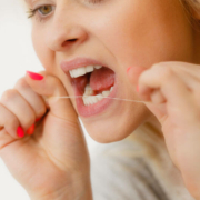 Eine Frau mit heller Haut und blondem Haar verwendet Zahnseide für ihre Zahnpflege. Sie fokussiert sich auf die gründliche Reinigung der Zahnzwischenräume. Ihre Mimik zeigt Konzentration und die Wichtigkeit der Mundhygiene. Die Zahnseide deutet auf die Empfehlung hin, ergänzend zu einer Schallzahnbürste, für optimale Sauberkeit und Gesunderhaltung der Zähne zu sorgen.